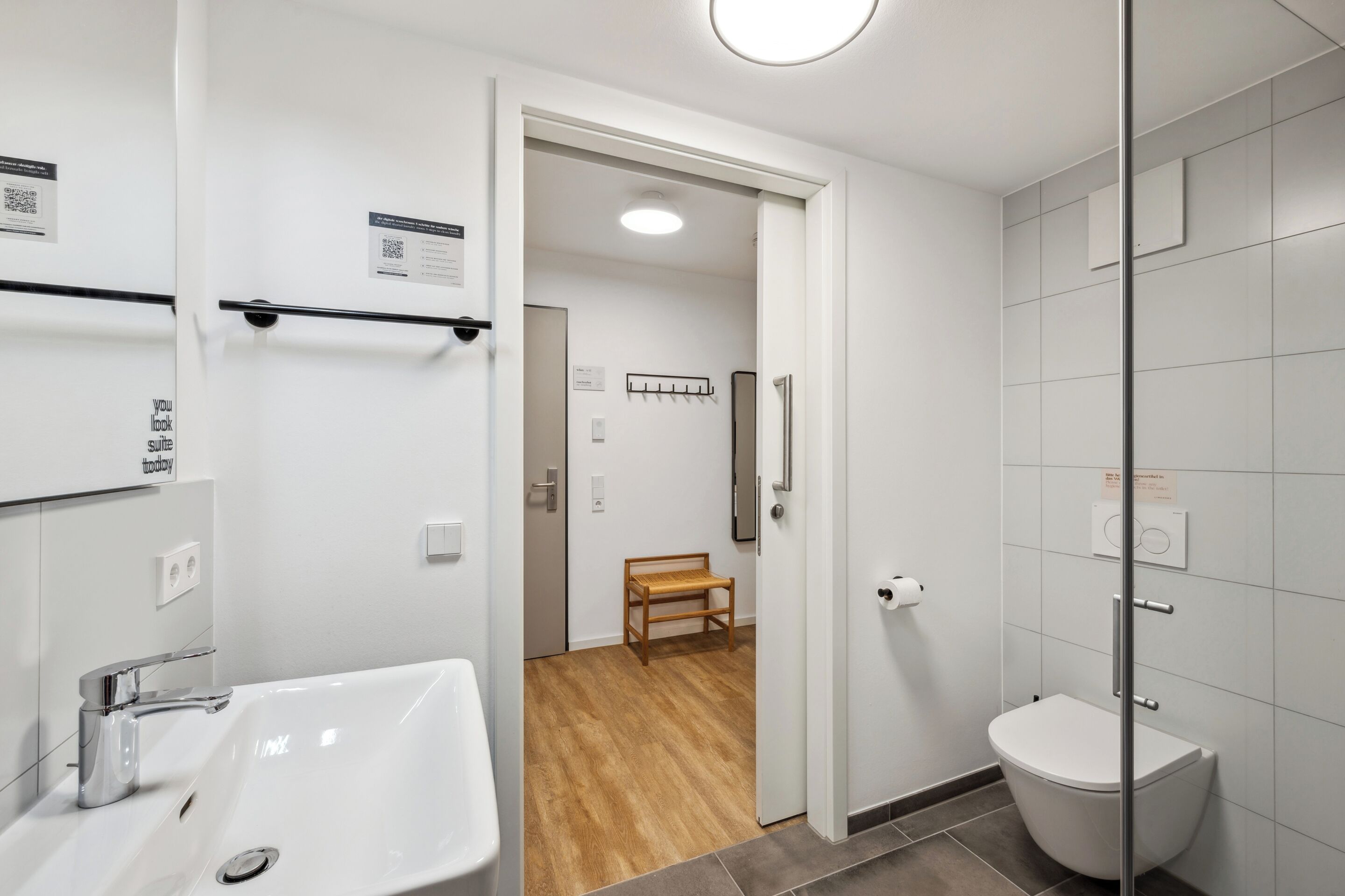 Une salle de bain moderne avec un lavabo blanc, des toilettes et un espace douche. On aperçoit un tabouret en bois à l’arrière-plan.