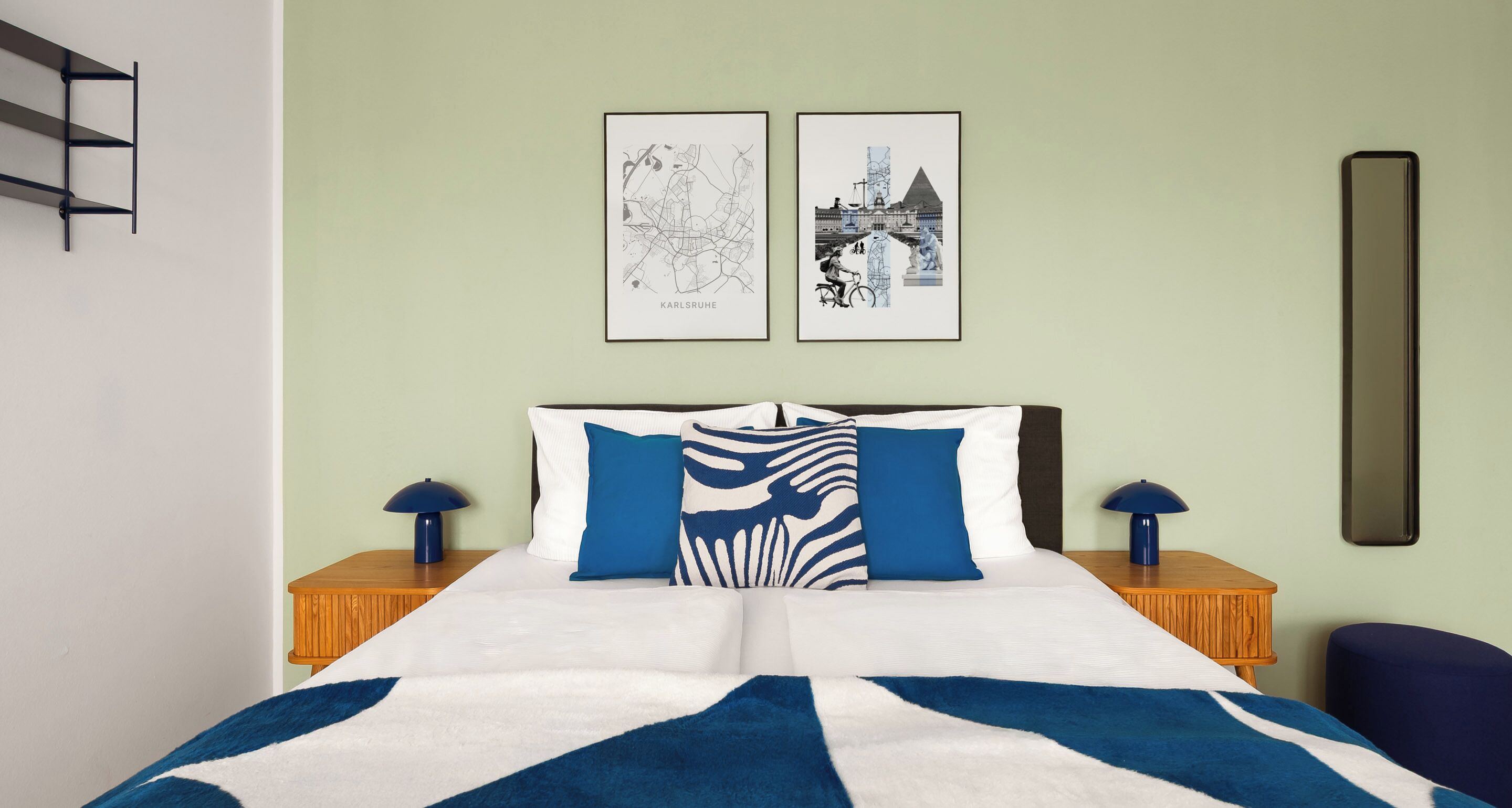 Una cama bien hecha con una colcha azul y blanca a rayas, dos almohadas y un cabecero. Hay dos cuadros enmarcados en la pared, una mesita de noche de madera con una lámpara y un espejo grande también en la pared.