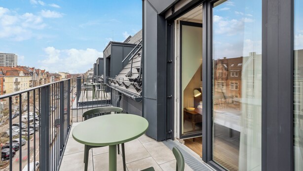Un balcon moderne avec une table et des chaises vertes, une porte vitrée donnant sur une pièce et une vue sur la ville.