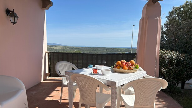A patio with a table set for two, white wicker chairs, and a basket of fruit on the table.