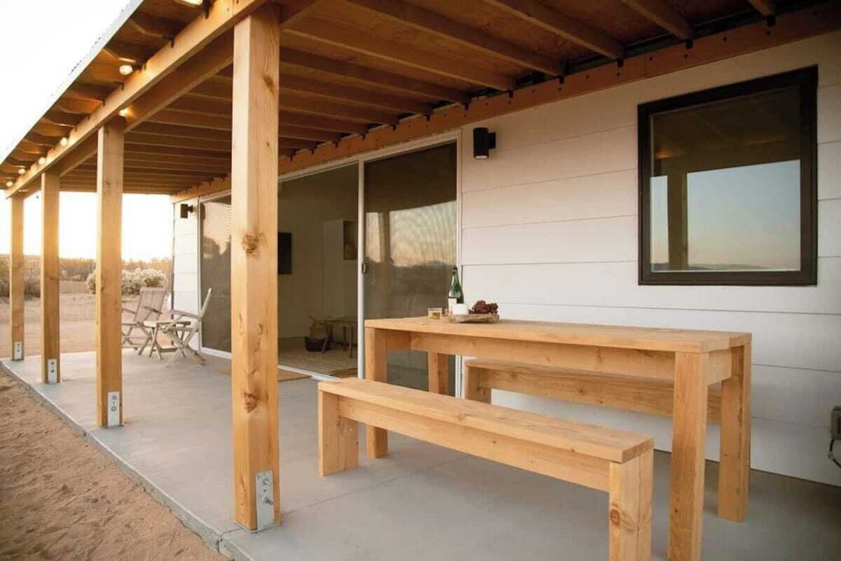 Eine Holzterrasse mit einer Bank und einem Tisch, einem Fenster und einer Tür, die in einen Innenbereich führt.