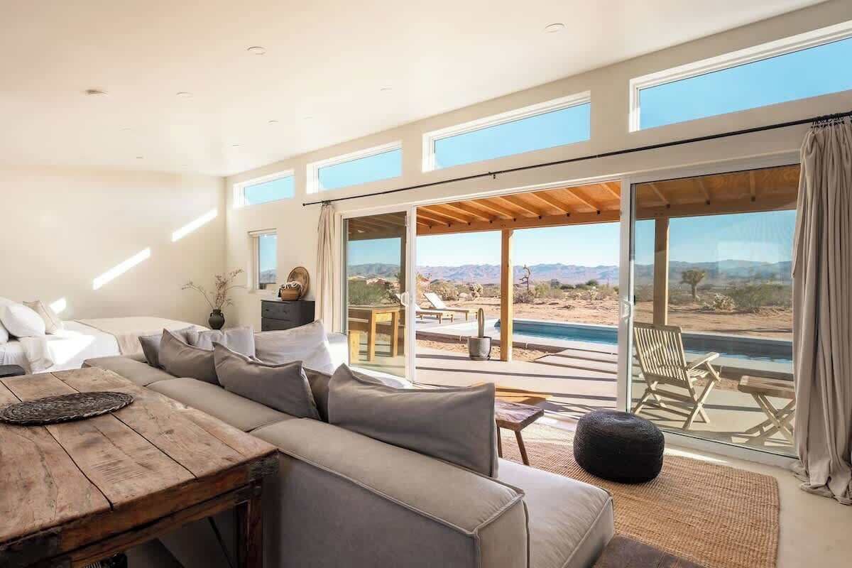 A modern living room with a large sofa, a wooden coffee table, and a view of a pool and mountains through large windows.