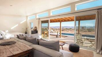 A modern living room with a large sofa, a wooden coffee table, and a view of a pool and mountains through large windows.