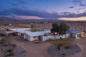 A modern house with a flat roof, large windows, and a garage, surrounded by a desert landscape.