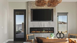 A modern living room with a flat-screen TV mounted above a fireplace, wicker pendant lights, and a view through a door to an outdoor area. - Free Bird • Pool, Spa, Pickleball, Outdoor Lvg Rm! (Joshua Tree)