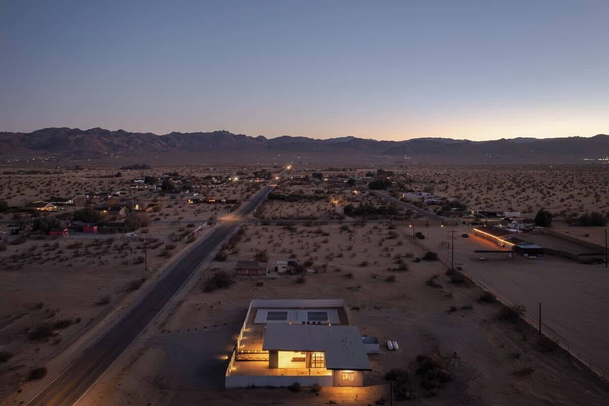 Vista aèria d'una vila al desert a l'hora del vespre, amb una carretera destacada que la travessa, envoltada de vegetació escassa i muntanyes llunyanes.