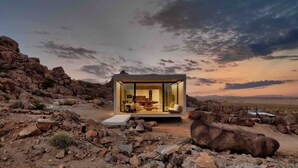 A modern, glass-walled structure in a rocky desert landscape with a sunset view. - Prism • Modern Luxury in Boulders, Pool & Hot Tub (Joshua Tree)