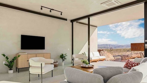 A modern living room with a sofa, a coffee table, and a TV mounted on the wall. There is a view of a desert landscape through large windows. - Prism • Modern Luxury in Boulders, Pool & Hot Tub (Joshua Tree)