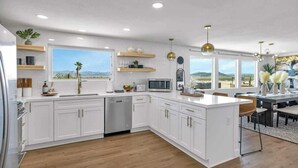 A modern kitchen with white cabinetry, a dishwasher, a microwave, and a large island. There are windows providing a view of the outdoors. - Winters Haven · 2.5 Acres, Spa, PingPong, SoakPool (Joshua Tree)