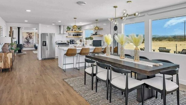 A modern kitchen with a central island, a dining area with a long table and chairs, and large windows offering a view of a desert landscape. - Winters Haven · 2.5 Acres, Spa, PingPong, SoakPool (Joshua Tree)