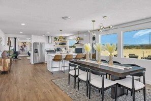 Una cocina moderna con una isla central, una zona de comedor con una mesa larga y sillas, y ventanas amplias que ofrecen vistas a un paisaje desértico.