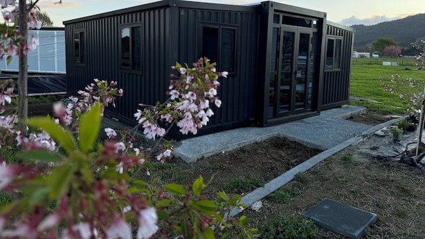 Un bâtiment moderne de couleur noire, doté de grandes fenêtres et d’une porte, entouré de verdure et surplombé d’un ciel dégagé.
