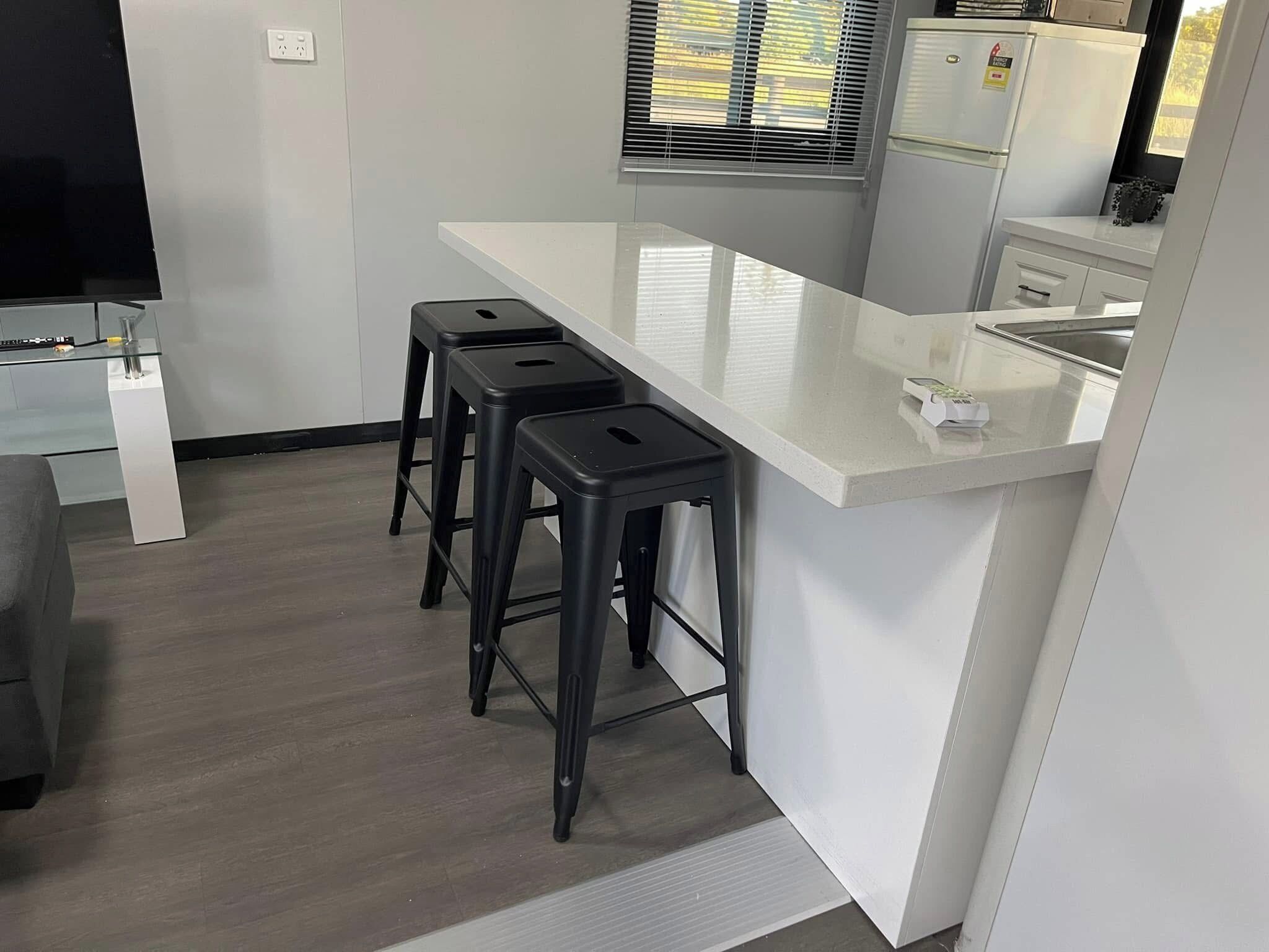 Een moderne keuken met een wit aanrecht, drie zwarte barstoelen, een roestvrijstalen spoelbak en een raam met zonwering.