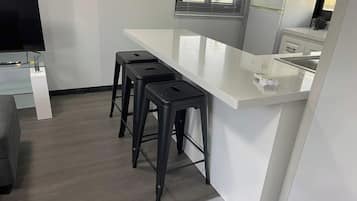 Een moderne keuken met een wit aanrecht, drie zwarte barstoelen, een roestvrijstalen spoelbak en een raam met zonwering.