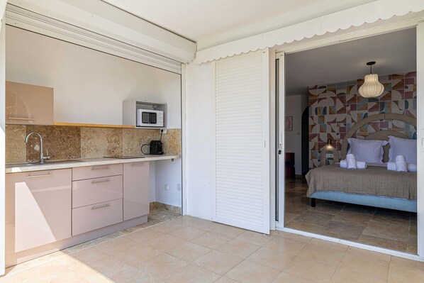 A compact bedroom with a bed, bedside table, and a patterned wall. Adjacent is a kitchenette with a microwave, coffee maker, and a small countertop.