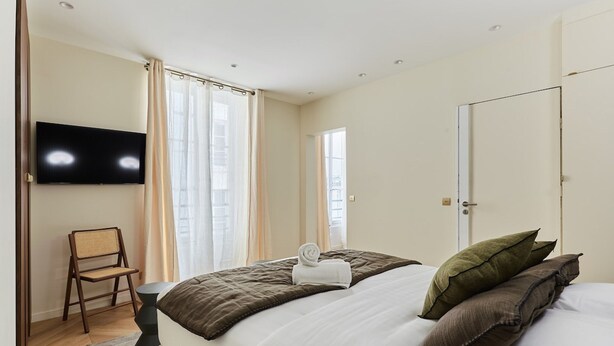 Habitación de hotel con cama, televisor de pantalla plana en la pared, una silla y ventana con cortinas.