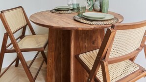 Una mesa de comedor redonda de madera con sillas tejidas, un florero con plantas secas y un pequeño florero verde sobre la mesa.