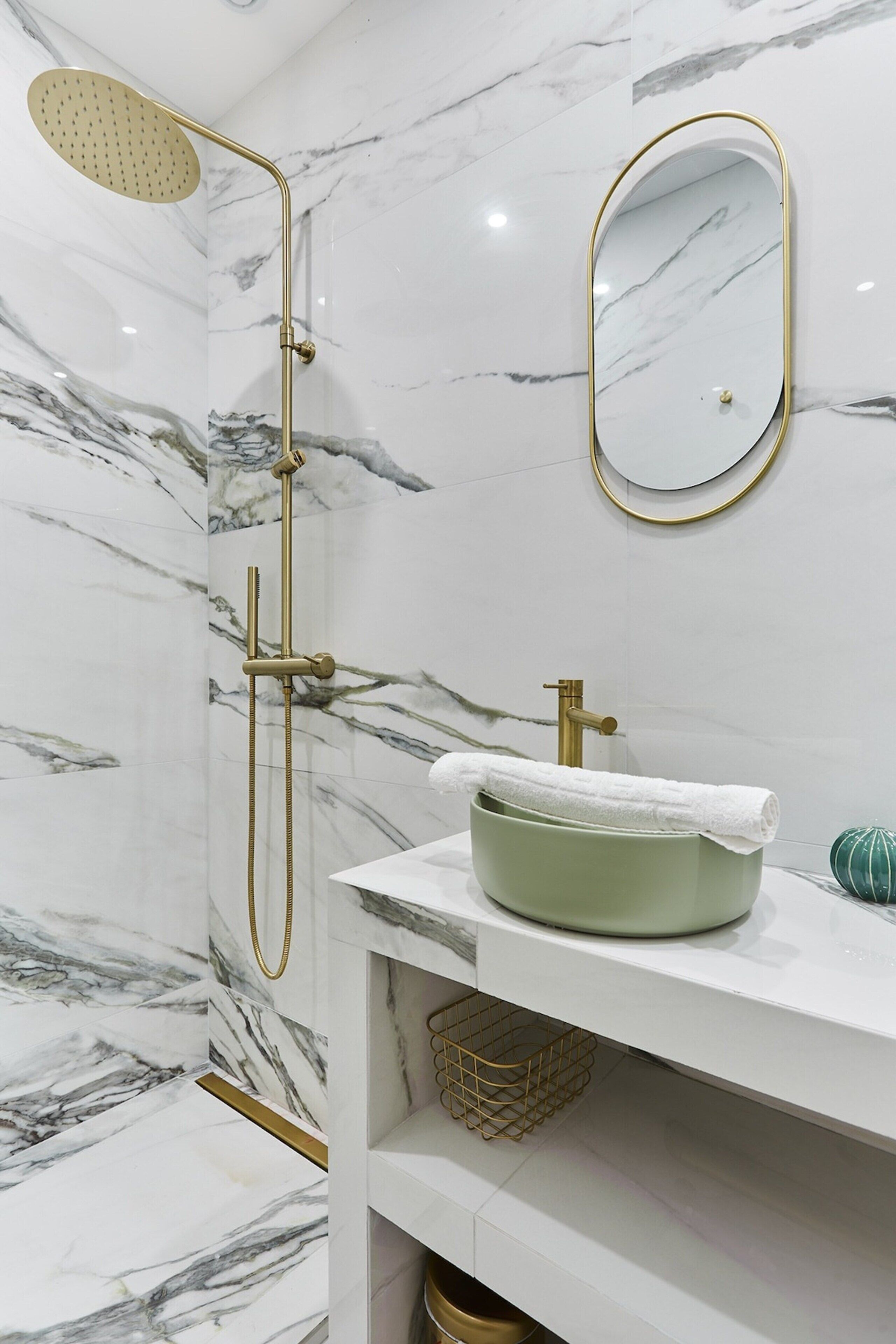A bathroom with a marble wall, a gold showerhead, a gold faucet, and a green sink.