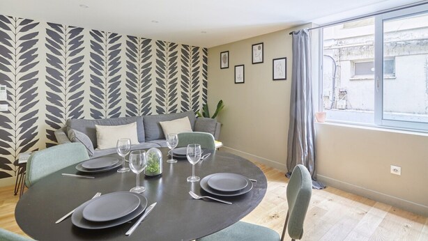 Een eetgedeelte met een ronde tafel voor vier personen, een bank en een groot raam met gordijnen.