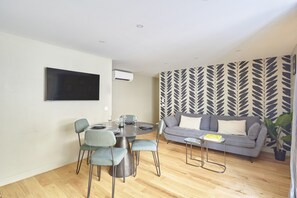Ein modernes Wohnzimmer mit grauem Sofa, einem Esstisch mit Stühlen und einem an der Wand befestigten Flachbildfernseher.