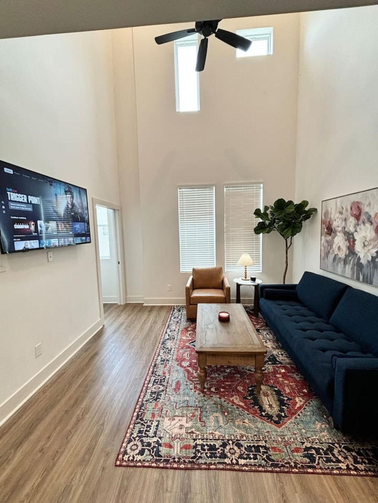 A spacious living room with a large flat-screen TV, a ceiling fan, a blue sofa, a coffee table, and a patterned rug.