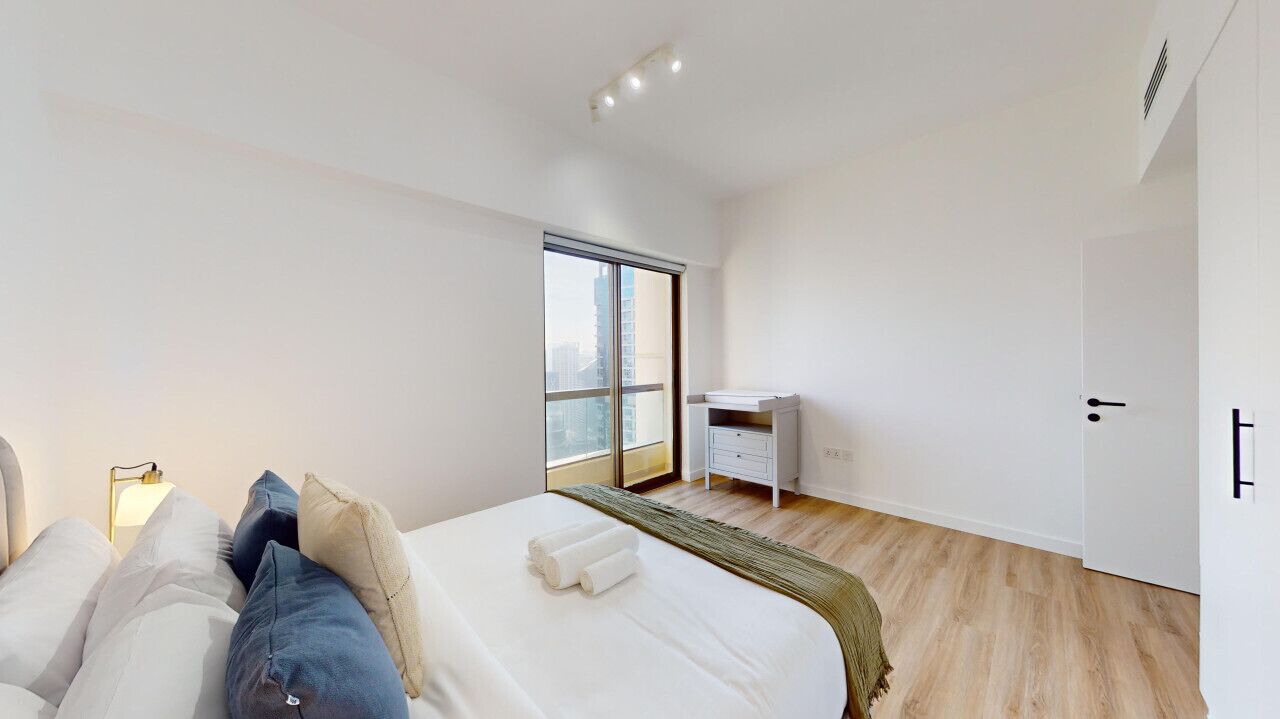 A modern hotel room with a large bed, a bedside table, a desk, and a wardrobe.