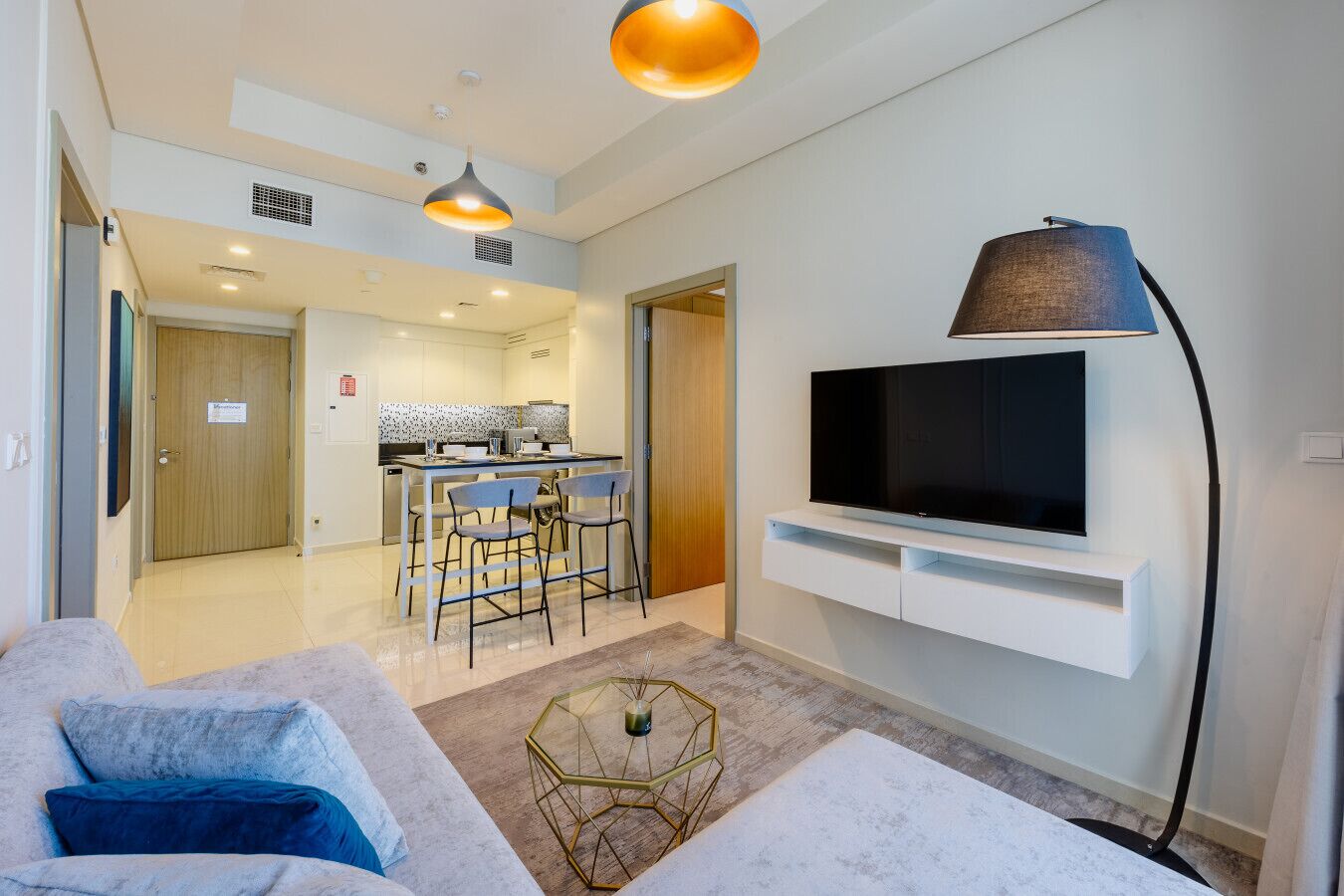 A modern living room with a sofa, a coffee table, a TV mounted on the wall, and a kitchen in the background.
