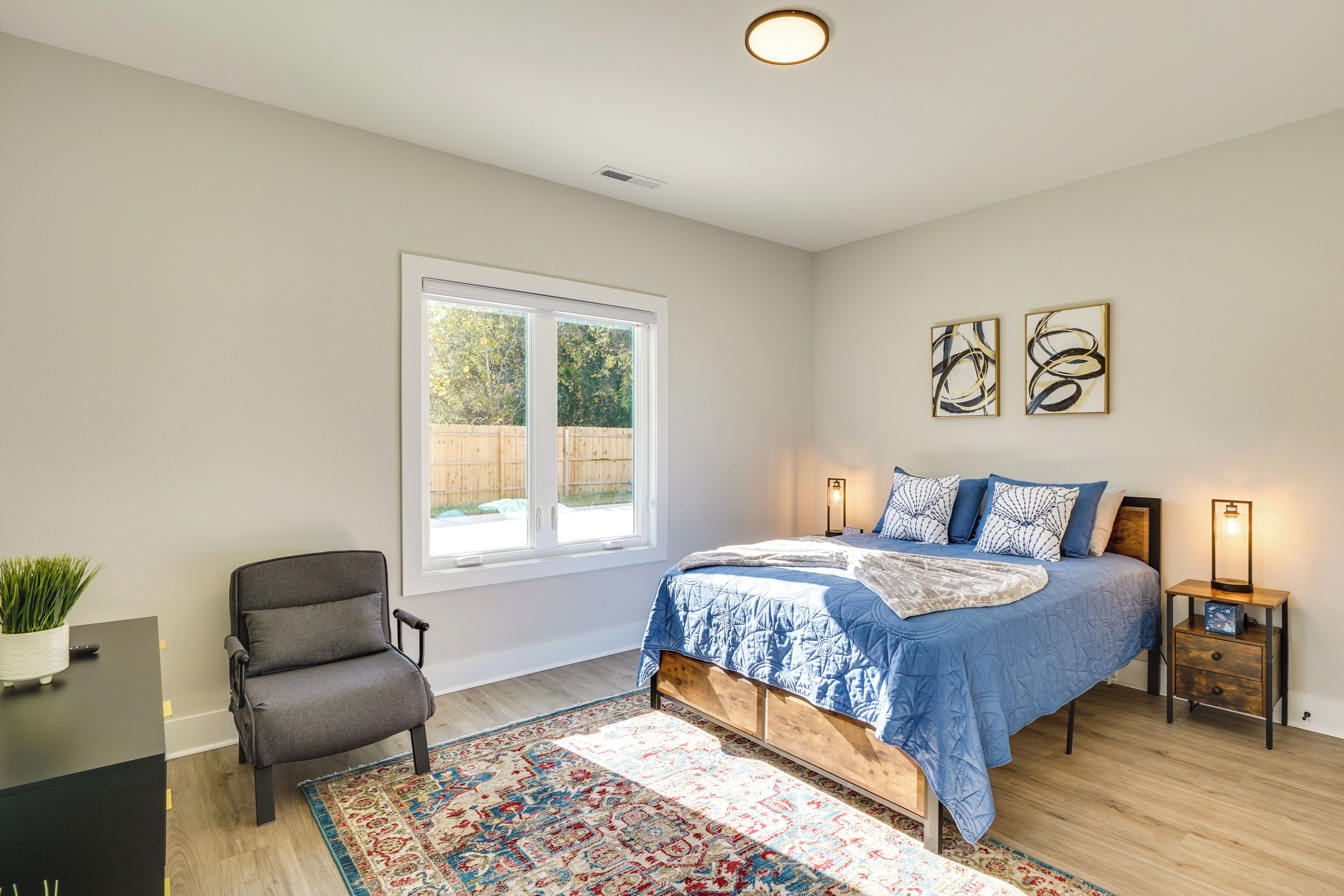 A bedroom with a large window, a bed with blue bedding, a chair, a bedside table, and a rug.