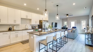 A modern kitchen with white cabinets, a large island with bar stools, stainless steel appliances, pendant lighting, and a dining area with a sofa and TV. - 2 Mi to Duke & 3 Mi to Downtown: Durham Townhome (Durham)