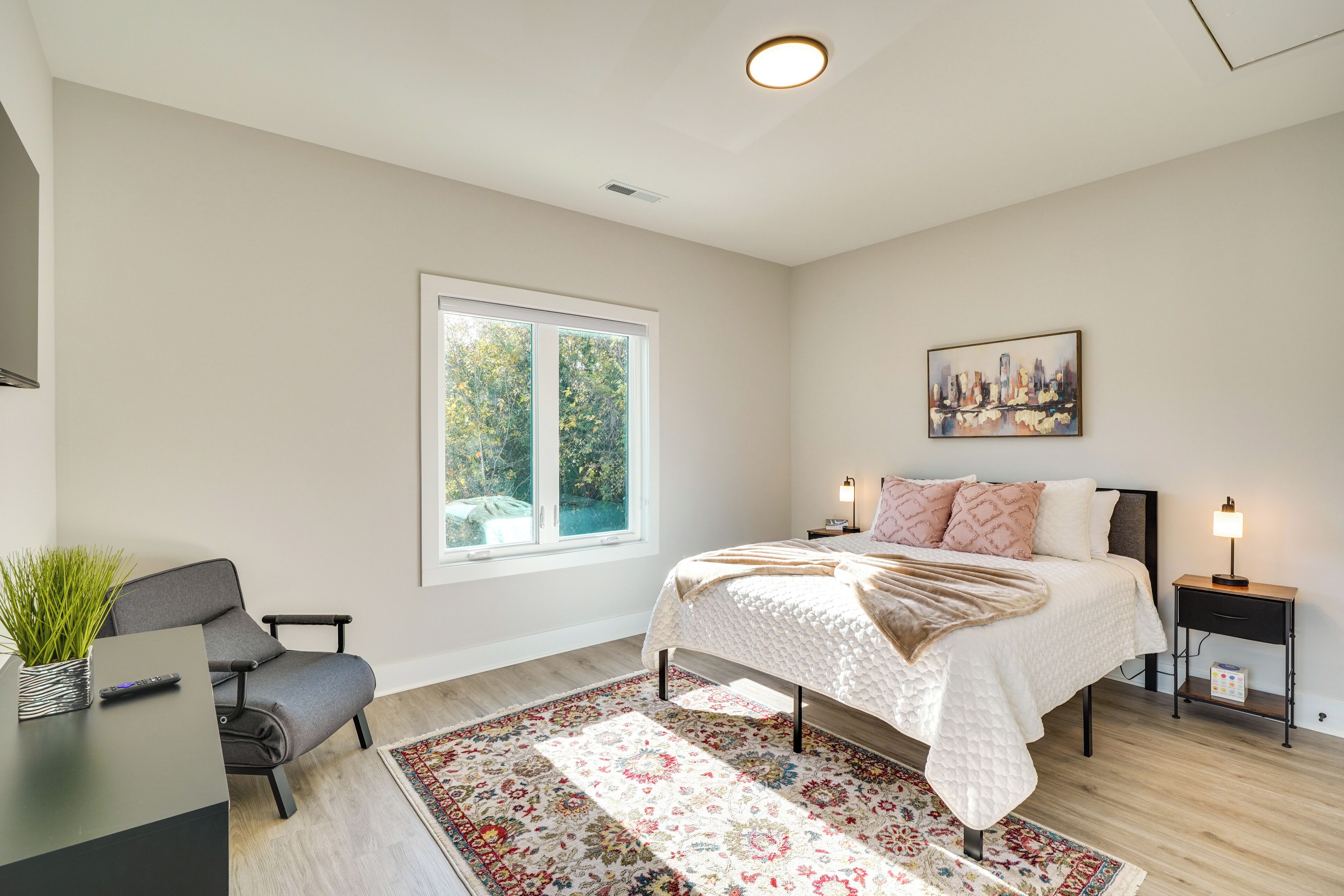 A bedroom with a large window, a bed with a white quilt, a nightstand with a lamp, and a chair with a plant.