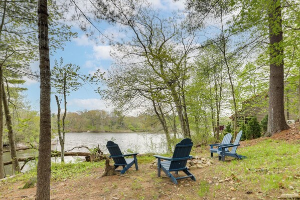 A lakeside setting with three Adirondack chairs facing the water, surrounded by trees and a clear sky.