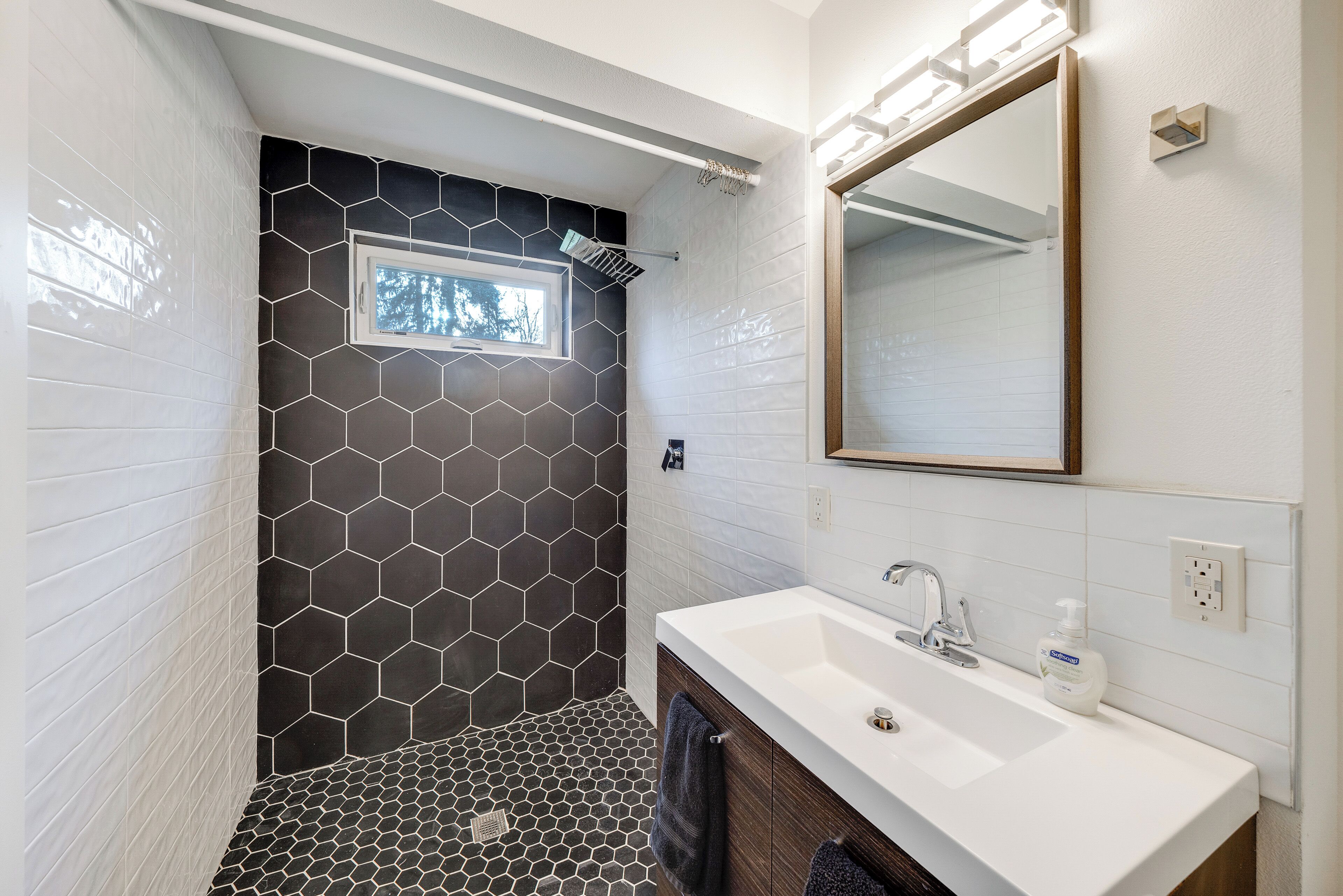 Une salle de bain moderne avec une douche carrelée de motifs hexagonaux, un lavabo blanc avec un miroir et une fenêtre située au-dessus de la douche.