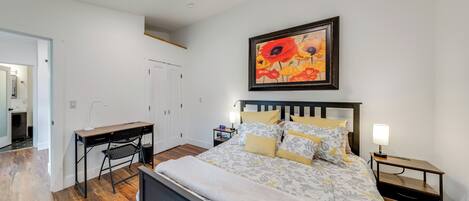 Une chambre à coucher avec un lit, un bureau, une chaise, une table de chevet et un tableau au mur.
