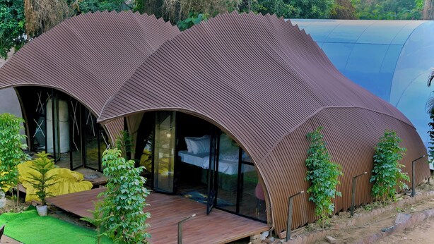 A modern, curved-roofed structure with a wooden deck and glass doors, surrounded by greenery. - Triveni Sangam Hotels And Resorts (Karchana)