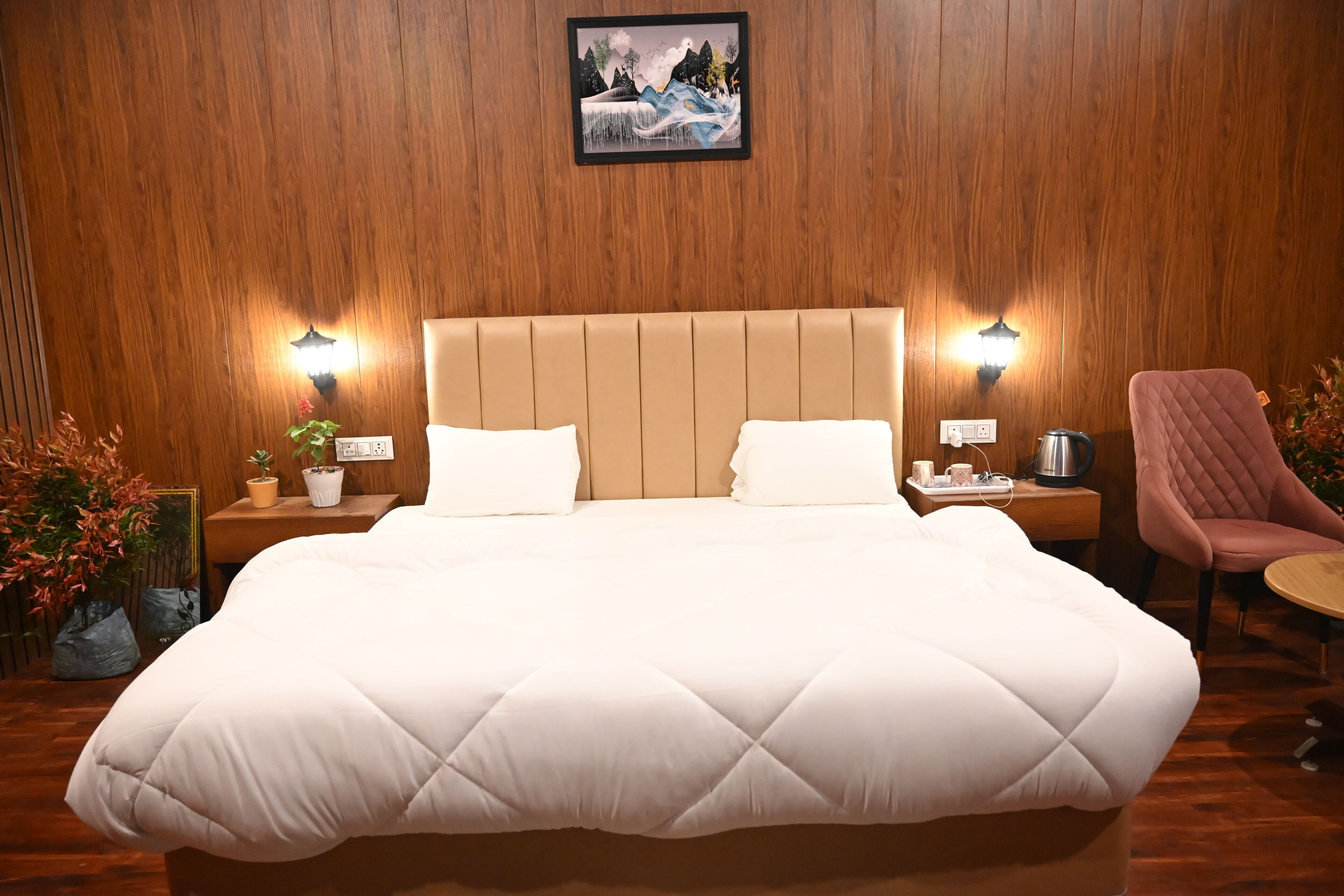 A hotel room with a large bed, wooden panel walls, two bedside lamps, a small table with a kettle, and a pink chair.
