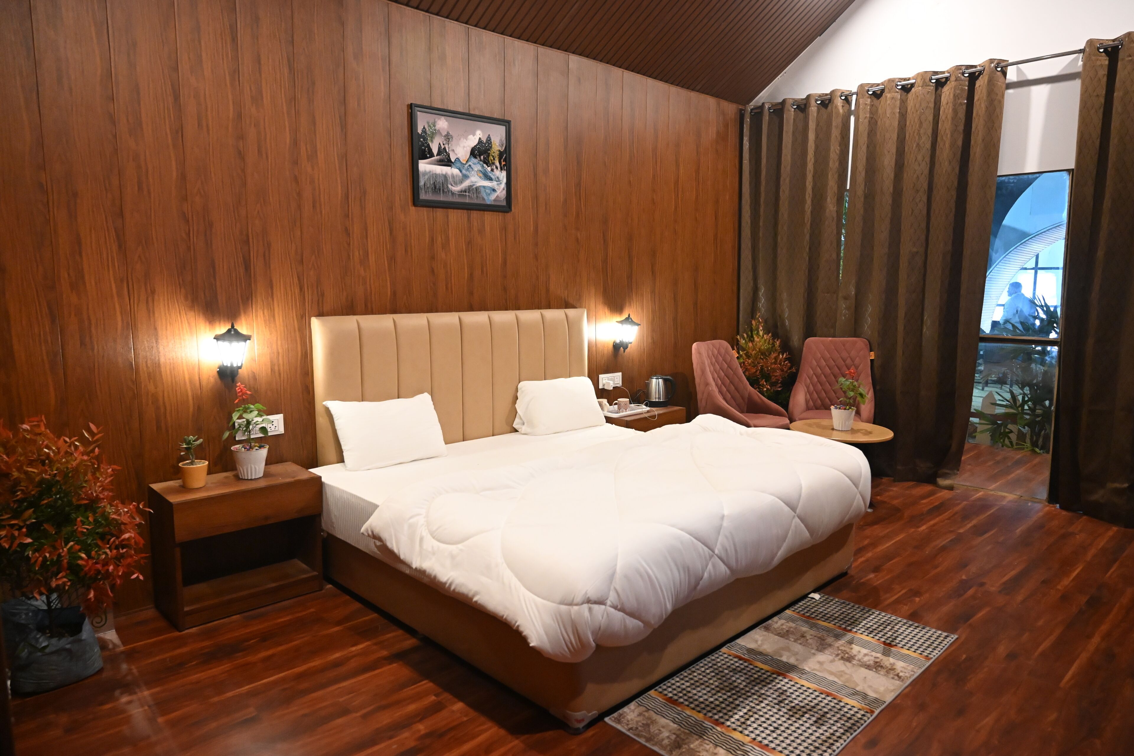 A spacious bedroom with a large bed, wooden walls, two bedside tables with lamps, a small table with a chair, and a view of the outdoors through a window.