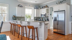 A modern kitchen with white cabinetry, a stainless steel refrigerator, and a bar area with wooden stools. - Oceanfront Home w/ Private Beach Access + Soaring Views by Tybee Cottages (Tybee Island)