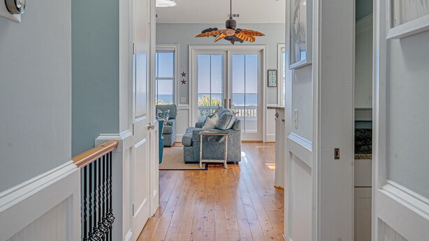 A hallway with wooden flooring, white walls, and a ceiling fan. There are two doors leading to a balcony with a view of the outdoors. - Oceanfront Home w/ Private Beach Access + Soaring Views by Tybee Cottages (Tybee Island)