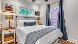 A bedroom with a bed, two bedside tables, a ceiling fan, a window with a view of trees, and a curtain.