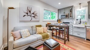A modern living room with a grey sofa, a coffee table, and a dining area with a wooden table and chairs. A kitchen with white cabinets and a microwave is visible in the background.