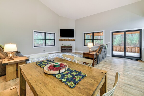 A spacious living room with a wooden dining table, a TV mounted on the wall, and large windows offering a view of the outdoors. - Brantingham Home: ATV & Snowmobile Trails On-site! (Brantingham)