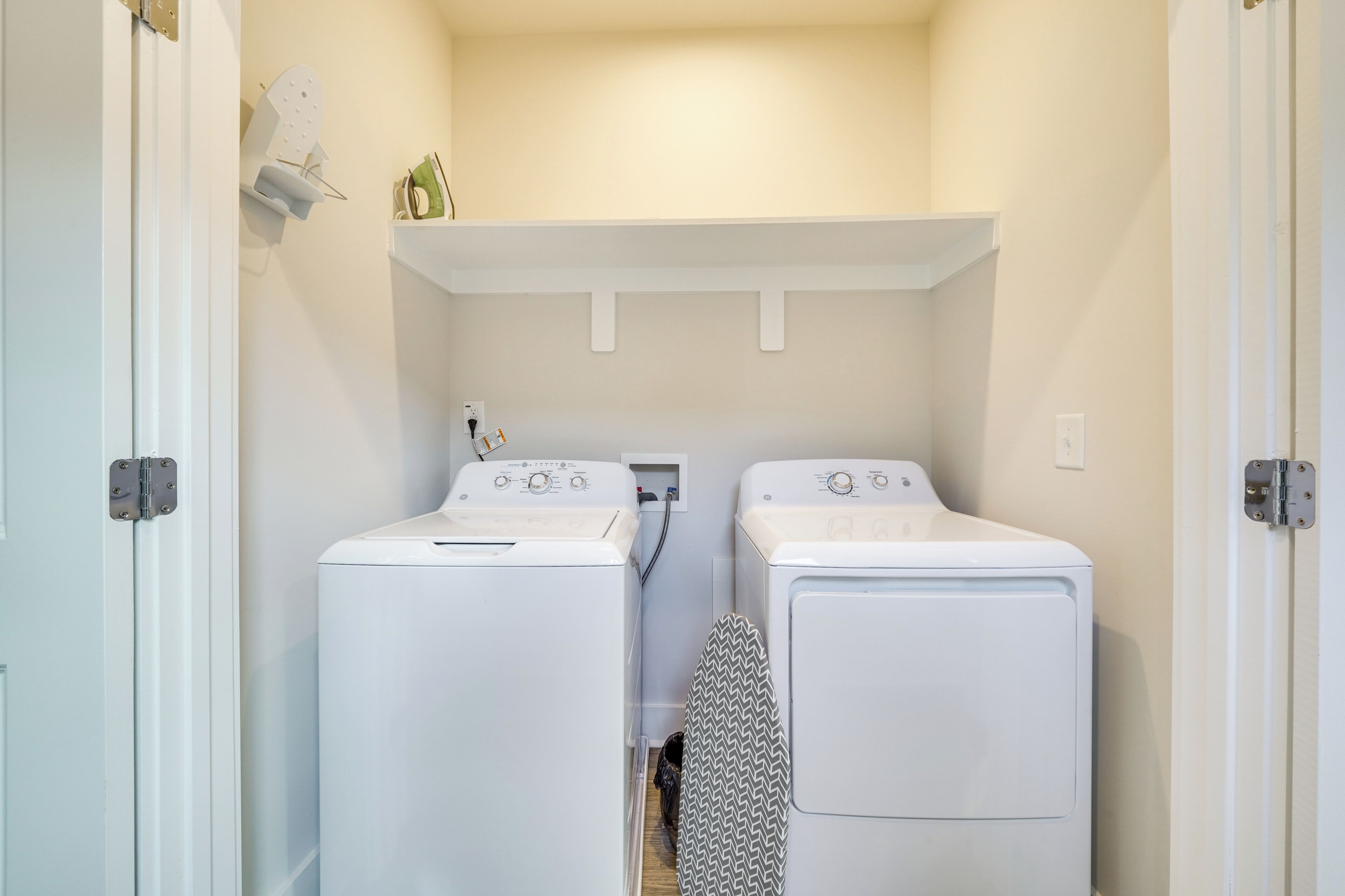 A laundry room with two white front-loading washing machines and a dryer, a small shelf above the machines, and a door on the left.