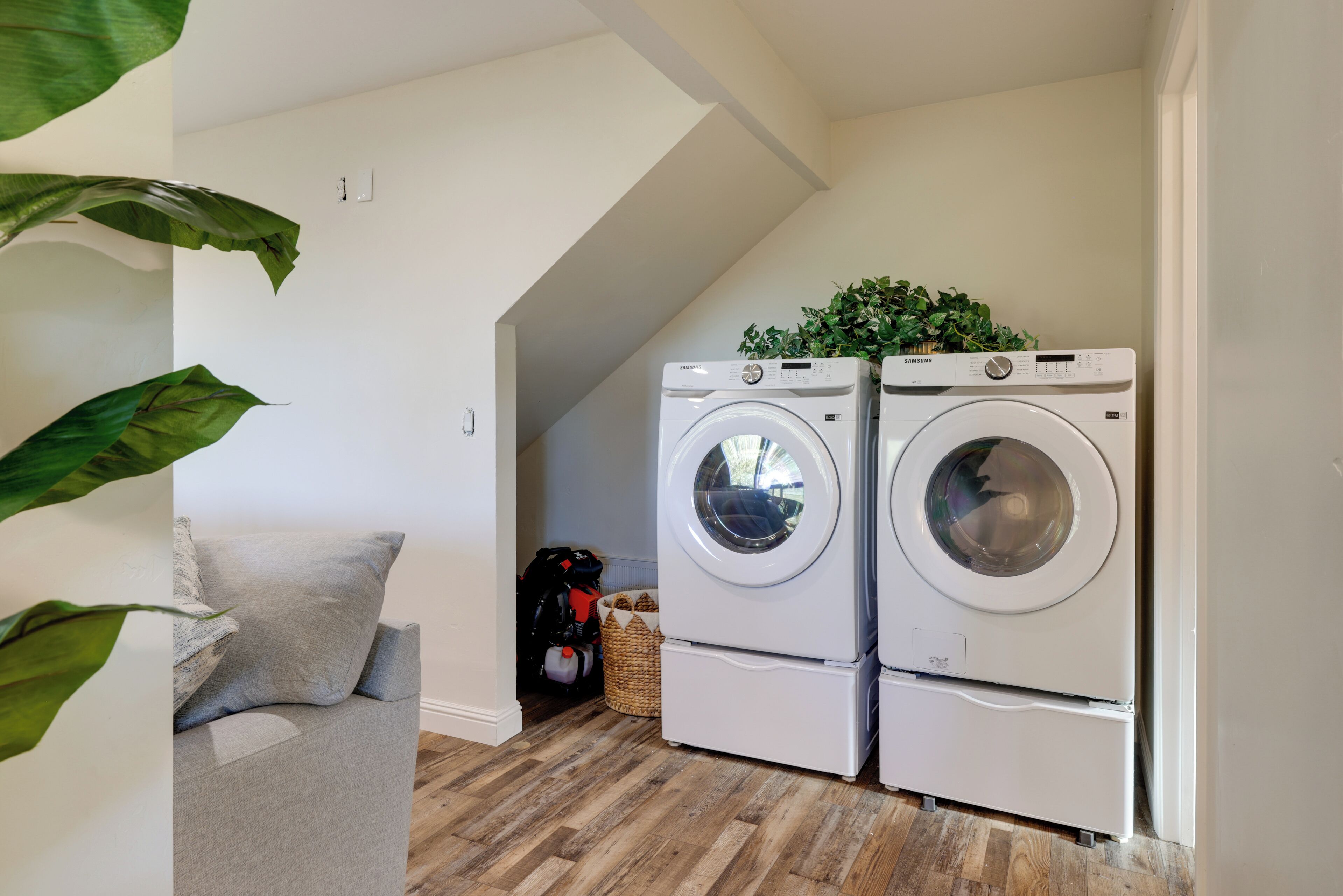 A laundry room with two stacked front-loading washing machines, a grey sofa, and a wooden floor.