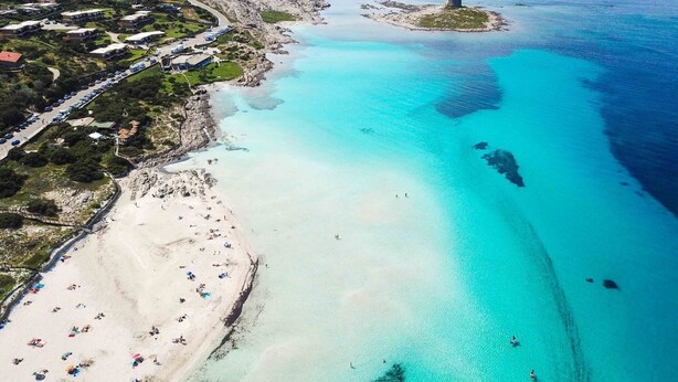 An aerial view of a coastal area with a sandy beach, clear turquoise waters, and a small island with a tower.