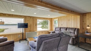 A cabin interior with a large window, a flat-screen TV, a sofa, and a table. - Waterfront Long Lake Retreat: Great for Families! (Sinclair)