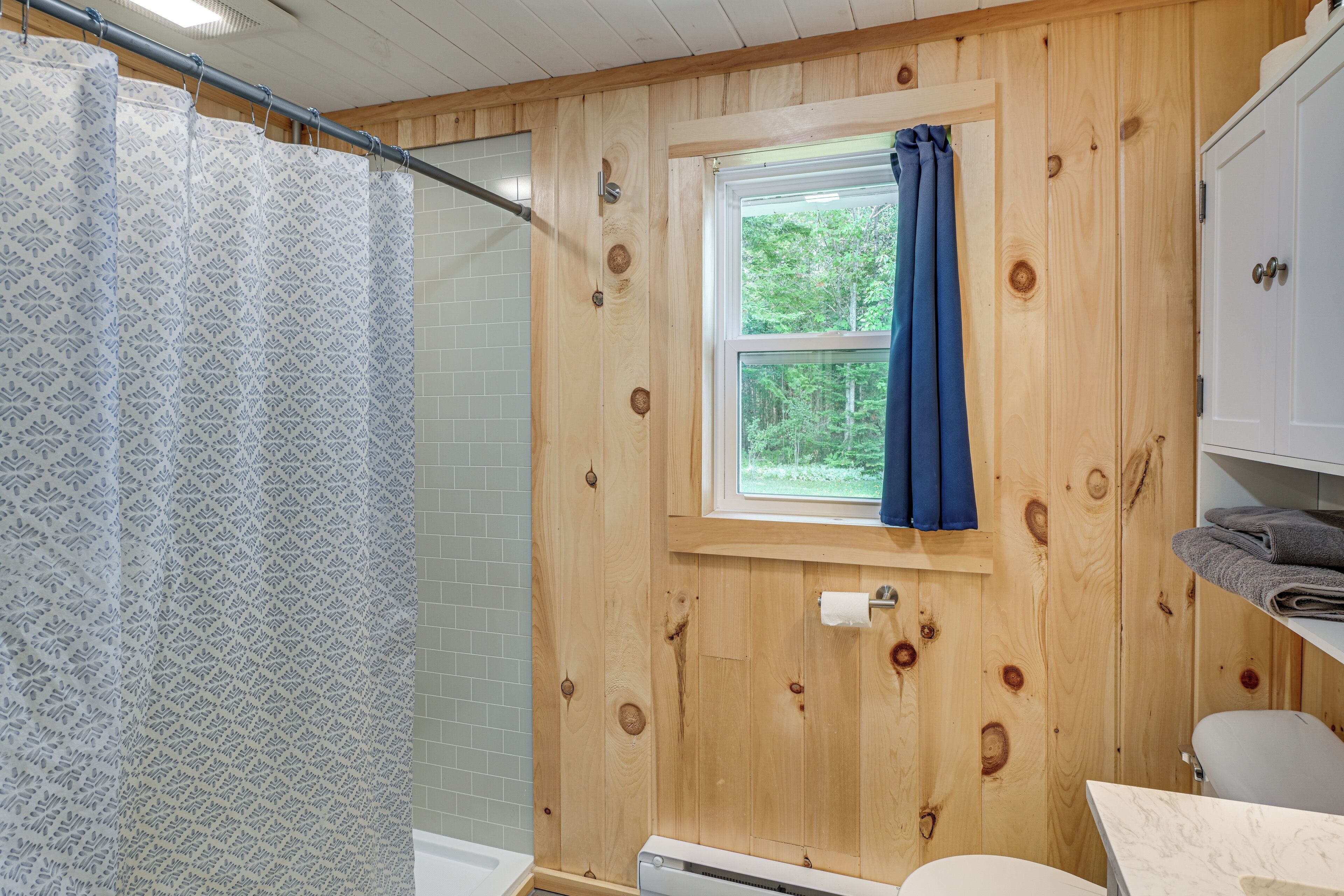 A bathroom with a shower curtain, a window with a curtain, a toilet, and a towel rack.