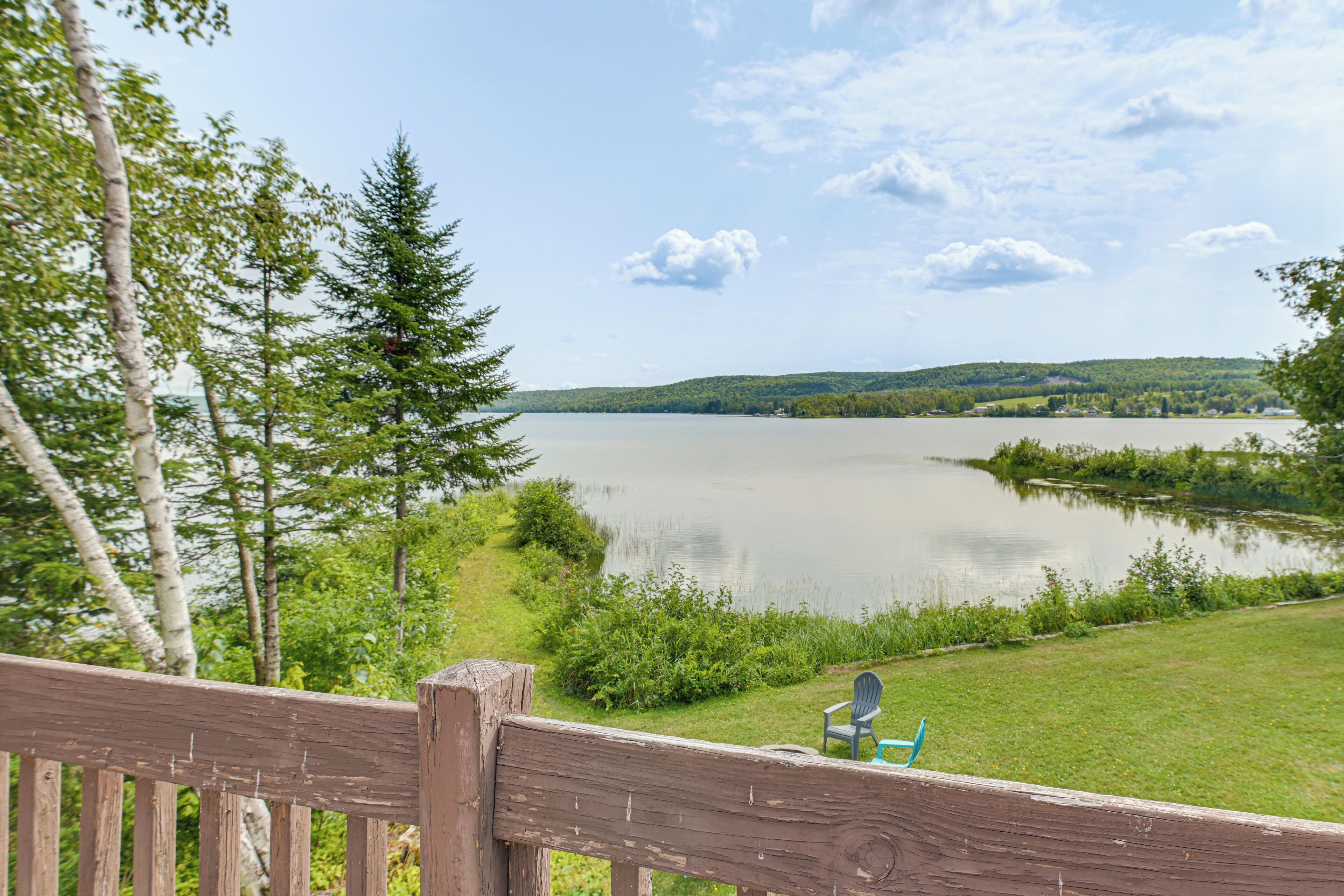 A lakeside view with a wooden deck and chairs, surrounded by greenery and a distant hill.