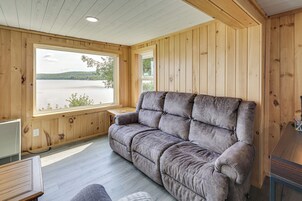 Interior de una cabaña de madera con un sofá gris, paredes de madera y una ventana con vistas a un lago.