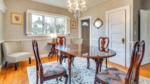 Uma sala de jantar com uma grande mesa de madeira, quatro cadeiras, um candelabro, uma janela com persianas e uma porta.