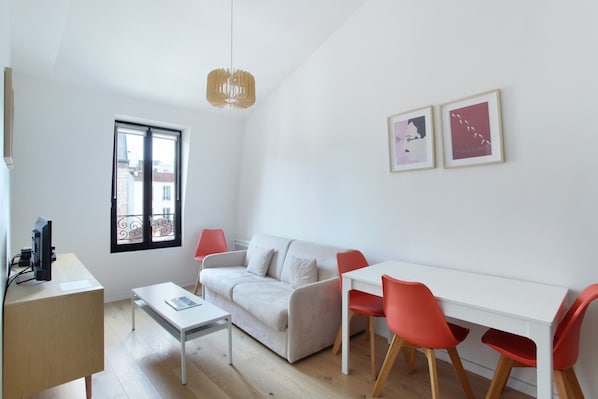 En moderne og lys stue med en hvid sofa, et lille bord og et spiseområde med røde stole. Der er et vindue med udsigt til bygninger, og to billeder hænger på væggen.
